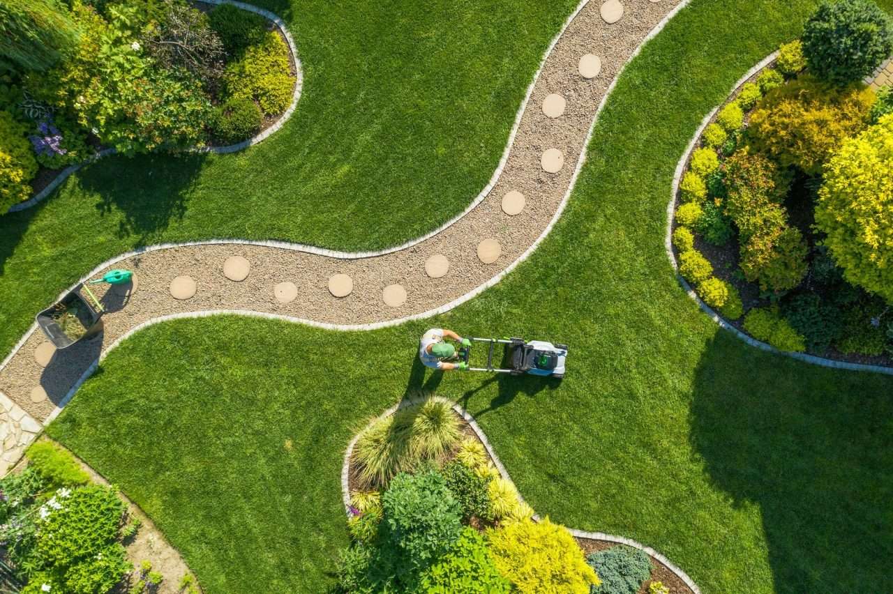 Caucasian gardener mowing a lush green lawn with a lawnmower, surrounded by well-maintained flower beds and garden pathways, illustrating professional landscaping services for lawn improvement and top dressing.