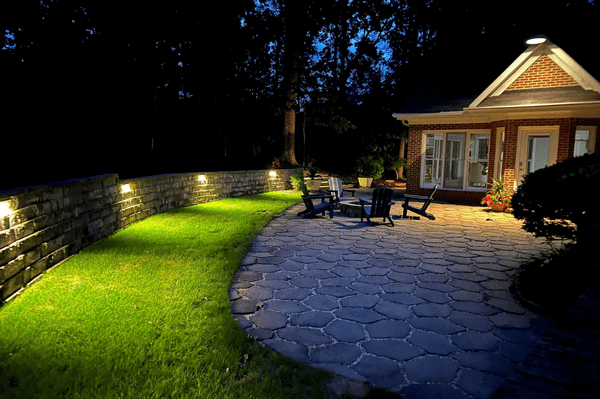 Landscape lighting illuminating a stone patio and grassy area beside a brick house at night, featuring outdoor seating and a well-maintained lawn, emphasizing aesthetic appeal for residential landscaping in Greenville, SC.