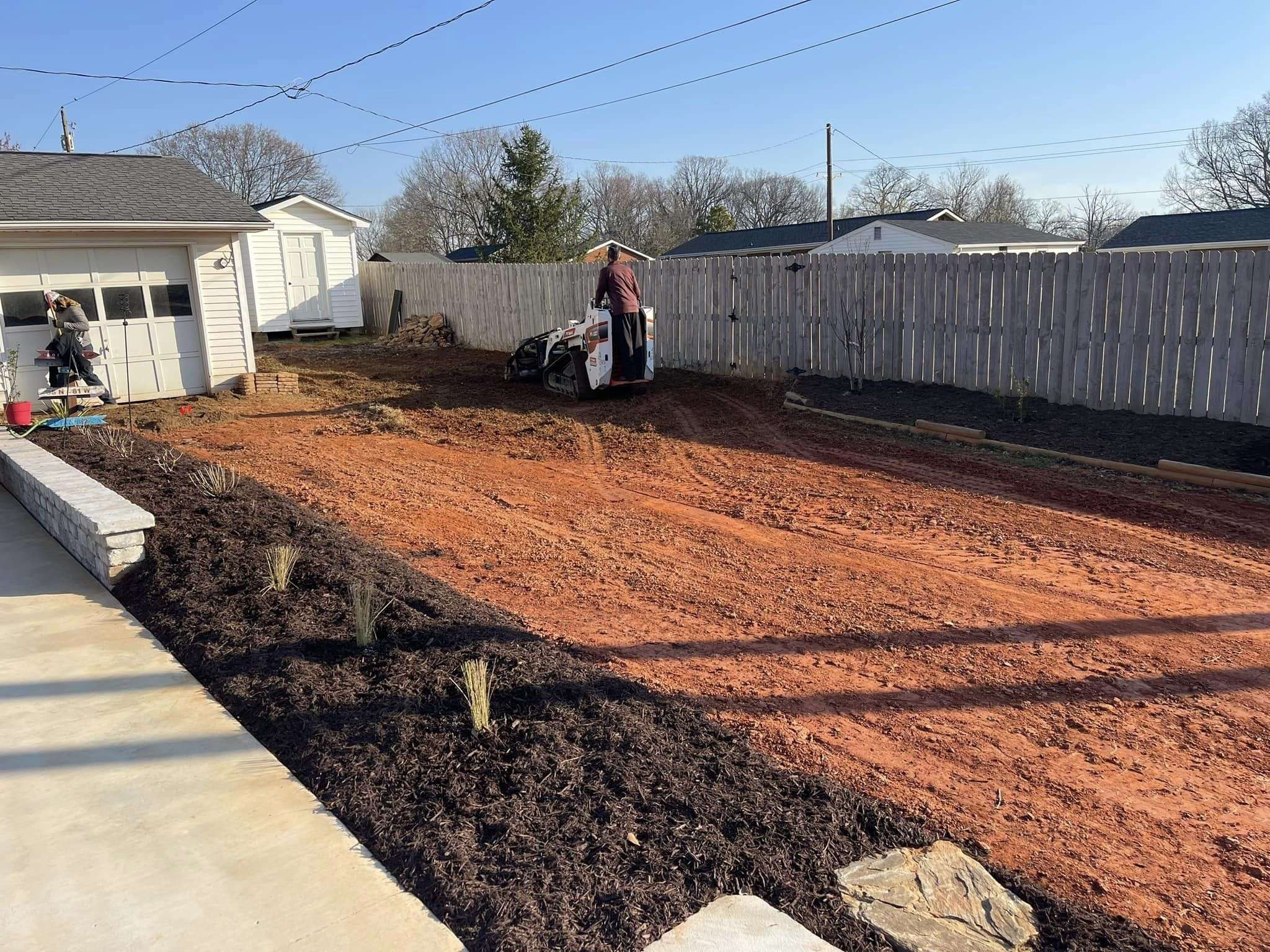 Soil preparation for sod installation, featuring a worker operating machinery on a freshly cleared yard with mulch and plants in the foreground.