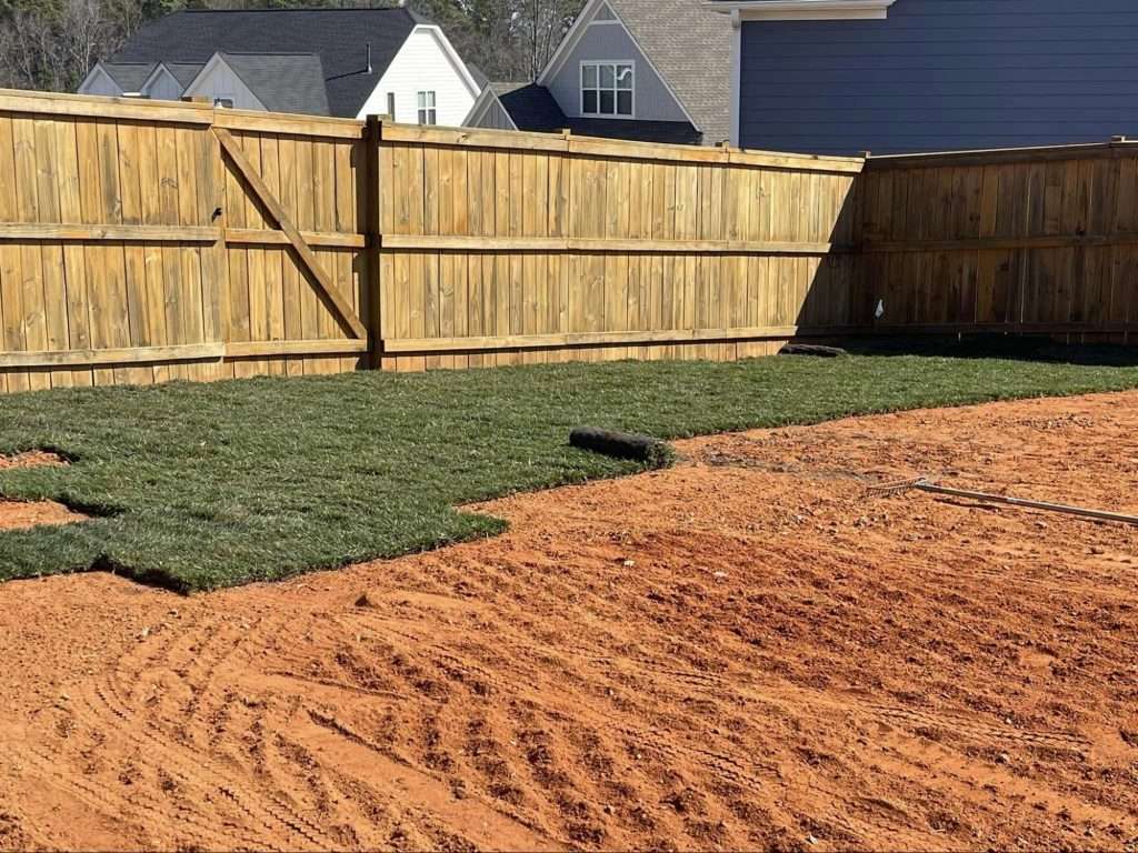 Freshly laid sod pieces in a backyard with visible red soil and a wooden fence