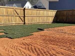 Newly installed sod in a yard with a wooden fence, showcasing a transition from bare soil to lush green grass, highlighting the importance of sod installation for landscaping.