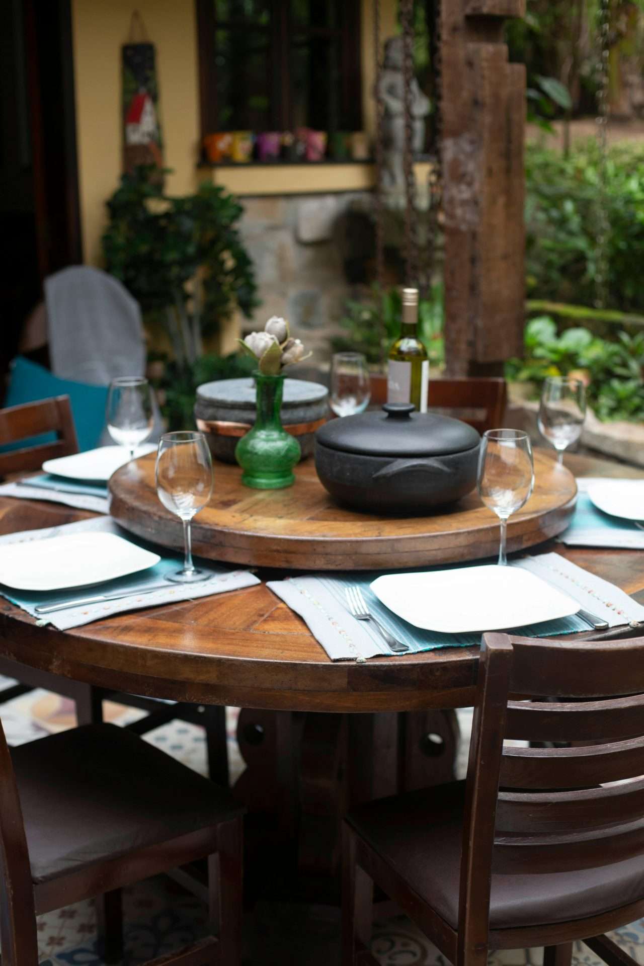 Outdoor dining table set for relaxation, featuring elegant plates, wine glasses, a decorative vase with flowers, and a black cooking pot, surrounded by a lush garden setting, highlighting the inviting atmosphere for alfresco dining experiences.