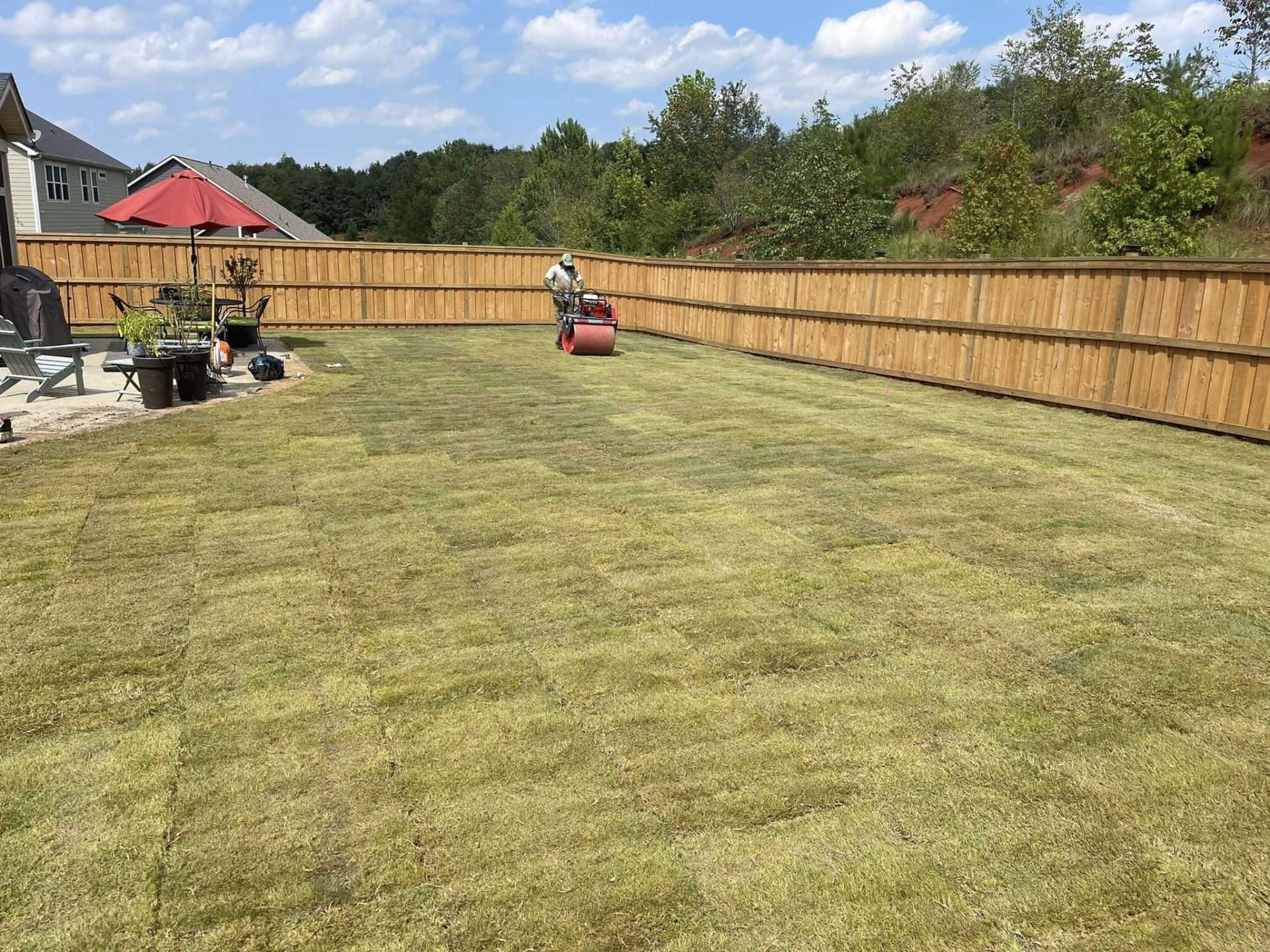 Person laying down freshly installed sod in a residential backyard in Greenville, SC, with a wooden fence and patio furniture visible.